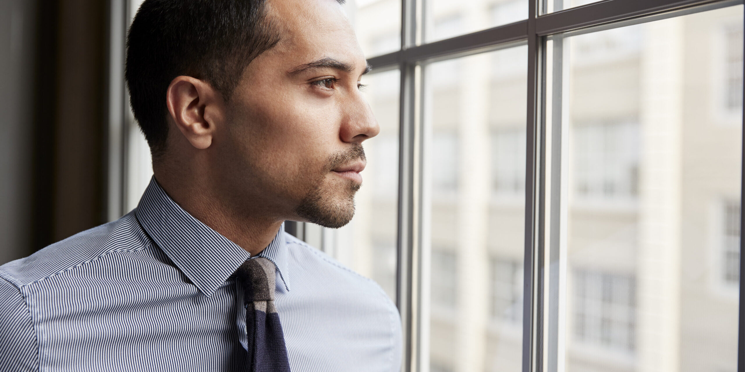 Corporate professional trapped by paycheck addiction looking out office window contemplating freedom