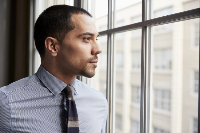 Corporate professional trapped by paycheck addiction looking out office window contemplating freedom