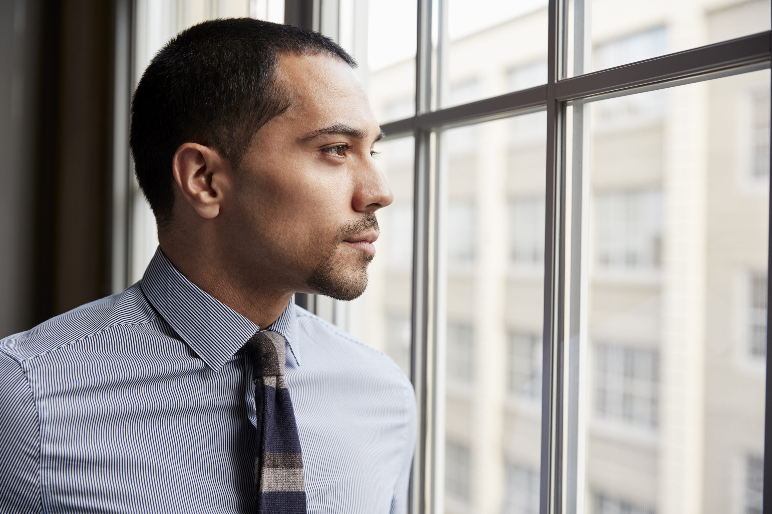Corporate professional trapped by paycheck addiction looking out office window contemplating freedom